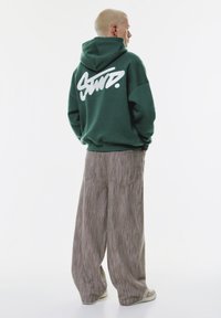 Person mit kurzen blonden Haaren, der einen dunkelgrünen Kapuzenpullover mit weißer Schrift und weiten grauen Hosen trägt und seitlich steht.
