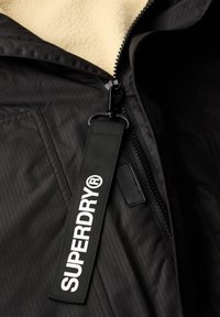 Chaqueta de tela negra a rayas con un forro de felpa color crema. Presenta una cremallera, un broche a presión y una etiqueta negra prominente con "SUPERDRY" en blanco.