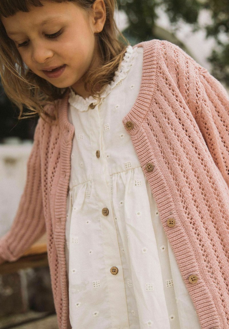 Ragazza giovane con vestito bianco in sangallo e cardigan rosa lavorato a maglia con bottoni in legno guarda in basso all'aperto.