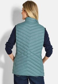 Helle tealfarbene, isolierte Weste mit einem Zickzackmuster und hohem Kragen, getragen über einem navyblauen Langarmhemd, das ein lässiges Design zeigt.