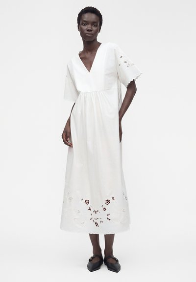 Femme portant une robe midi blanche avec col en V, manches courtes ajourées, détails floraux découpés à l'ourlet et ballerines noires pointues.