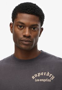 Grå t-shirt lavet af blødt stof, med et buet logo i lys beige tekst på venstre side. Rund hals, korte ærmer.