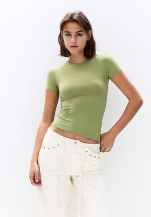 Korte mouwen, strakke top in lichtgroen, gemaakt van rekbaar materiaal. Gecombineerd met losse, witte broek met zilveren studs.