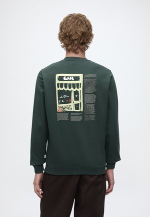 Personne aux cheveux bouclés portant un sweat-shirt vert foncé avec une illustration de devanture de café et un texte au dos.