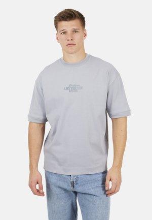 Giovane uomo che indossa una felpa a maniche corte grigio chiaro con la scritta "Amsterdam EST. 1972" e jeans azzurri, in piedi davanti a uno sfondo bianco.