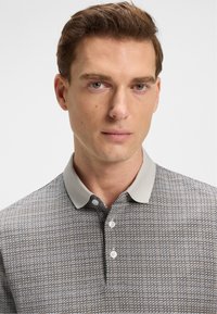 Hombre con cabello corto castaño que lleva una camiseta polo de color gris con un cuello liso de un gris claro, mirando de frente sobre un fondo blanco.