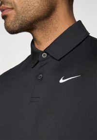 Polo noir avec un col, deux boutons et un logo Nike en blanc sur la poitrine gauche. Texture lisse et design ajusté.