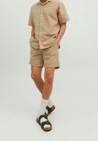 Personne portant une chemise beige à manches courtes et un short assorti, des chaussettes blanches et des sandales noires à boucle, se tenant les mains dans les poches.