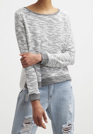 Pull en gris clair avec un motif en vagues texturé, encolure ras du cou et poignets en gris foncé contrastants. Porté par-dessus un jean bleu clair usé.