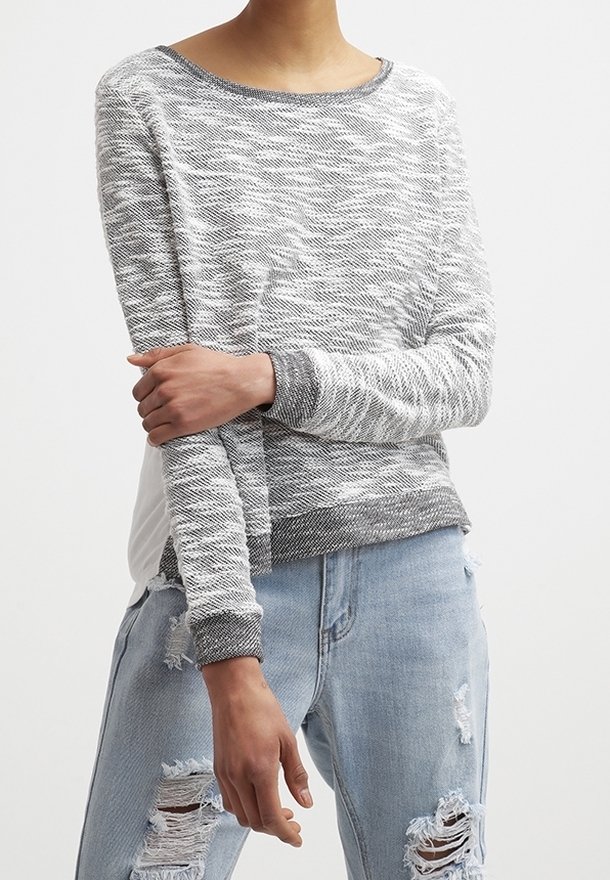 Hellgrauer Pullover mit einem strukturierten Wellenmuster, Rundhalsausschnitt und kontrastierenden dunkelgrauen Bündchen. Getragen über zerrissenen hellblauen Jeans.