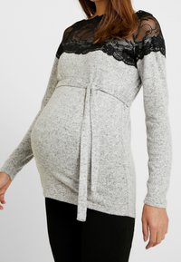 Haut de maternité gris à manches longues, avec un empiècement en dentelle noire et un détail de taille nouée, confectionné dans un tissu doux et texturé.