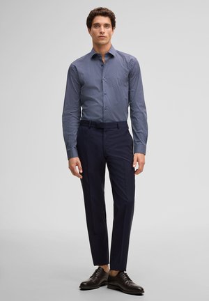 Jonge man staand, gekleed in een blauw overhemd met patroon, donkere aansluitende broek en zwarte leren nette schoenen.