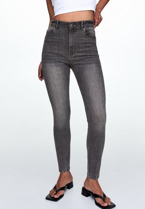 Osoba ubrana w wyblakłe czarne obcisłe dżinsy i czarne sandały na obcasie z odkrytymi palcami na jednolitym jasnym tle.