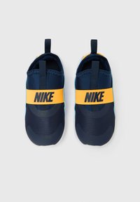 Paire de baskets Nike bleu marine à enfiler avec des sangles élastiques jaunes au milieu du pied, vues de dessus sur un fond clair.