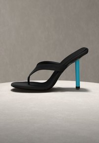 Melna augstpapēžu sandale ar pirkstu siksniņu un gaiši zilu stiletto papēdi uz gaišas apaļas virsmas pret vienkāršu pelēku fonu.