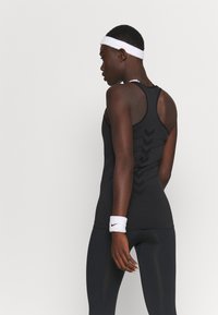 Woman in black athletic tank top and leggings, wearing white headband and wristband, facing sideways against plain white background.