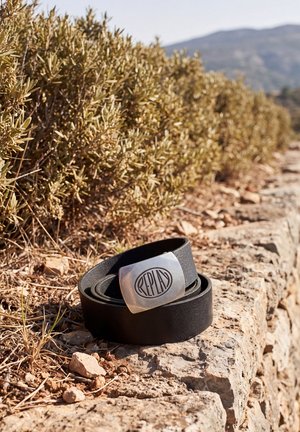 Un cinturón de cuero negro con una hebilla plateada de Replay reposa en un sendero pedregoso junto a unos arbustos secos. Al fondo, las montañas aparecen desenfocadas.