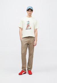 Κρεμ κο ντομάνικη μπλούζα με γραφικό σχέδιο και κείμενο "VOLCOM", σετ με μπεζ παντελόνι και κόκκινα αθλητικά με μαύρες λεπτομέρειες. Ανοιχτό μπλε καπέλο.