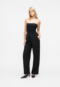 Νεαρή γυναίκα όρθια, φορώντας μαύρο τοπ χωρίς τιράντες, μαύρο φαρδύ παντελόνι και μαύρα παπούτσια με χαμηλό τακούνι, σε λευκό φόντο.