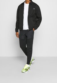 Černá bomber bunda přes bílé tričko, doplněná černými kalhotami s náprsními kapsami a jasnými, vzorovanými teniskami.