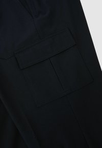 Gros plan sur un tissu noir montrant une poche rectangulaire avec un rabat et des coutures visibles sur la face avant d'un vêtement.