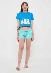 T-shirt blu corto con grafica di un sole bianco, abbinato a pantaloni corti azzurri e sandali bianchi con logo. Texture liscia, design casual.