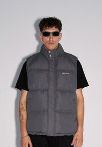 Szary kamizelka z wysokim kołnierzem, zapięciem na napy i kieszeniami po bokach. Nosi się ją na czarnej koszulce; model ma także na sobie okulary przeciwsłoneczne.