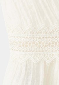 Tissu blanc avec une bande de dentelle horizontale présentant des motifs floraux et géométriques au-dessus d'un matériau froncé et texturé.