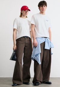 Δύο μοντέλα φορούν λευκά γραφικά μπλουζάκια και φαρδιά καφέ παντελόνια· το ένα κρατά μια μαύρη τσάντα και φορά κόκκινο καπέλο, ενώ το άλλο έχει μπλε πουκάμισο δεμένο στη μέση.