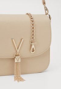 Borsa in sintetico beige con chiusura a pattina, tracolla in catena dorata e un tassello decorativo con un'emblema a forma di V dorata.