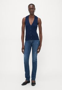 Ženska v brezrokavnem modrem zgornjem delu s gumbi, tesno prilegajočih iznakaženih modrih kavbojkah, črnih ravnih čevljih in uhani v obliki obroča, stoji na beli podlagi.