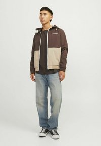 Chaqueta con capucha en bloques de color marrón y beige, con cierre de cremallera, puños elásticos, usada sobre una camisa oscura con vaqueros de color azul claro y zapatillas negras.