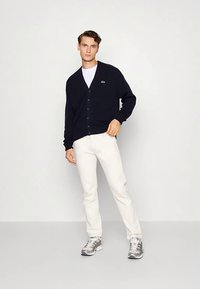 Marineblå cardigan med V-hals, knappelukking, ribbestrikkede mansjetter, båret over en hvit T-skjorte. Kombinert med lyse bukser og sølvfargede joggesko.