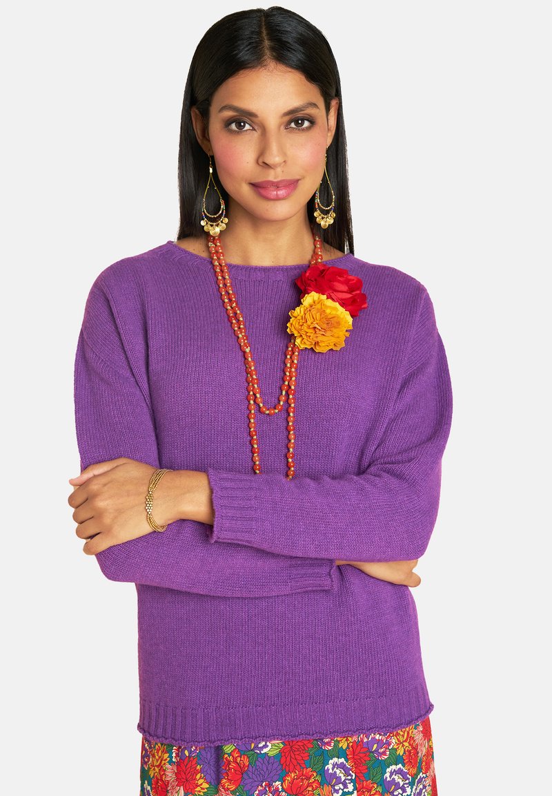 Frau mit dunklem Haar in einem lila Pullover, einem Blumenkleid, einer roten und gelben Blumenbrosche, einer roten Perlenkette, einem goldenen Armband und hängenden Ohrringen.