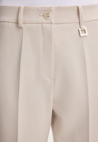 Pantaloni beige chiaro realizzati in tessuto liscio, dotati di piega frontale, tasca singola e un dettaglio in metallo quadrato argentato sulla cintura.