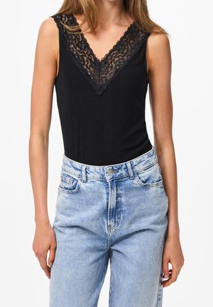 Kobieta ubrana w czarny, bezrękawnik z koronkowym dekoltem w kształcie litery V, zestawiony z wysokim stanem jasnoniebieskich jeansów.