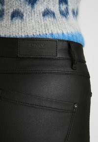 Pantalons en simili cuir noir avec une texture lisse, présentant des coutures surpiquées, des boutons métalliques et une étiquette noire de la marque au niveau de la taille.