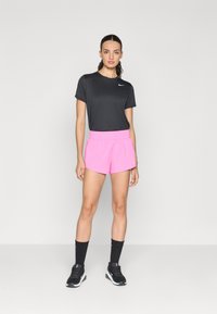 Mujer de pie contra un fondo blanco que lleva una camiseta negra de Nike, pantalones cortos rosa brillante, calcetas negras y zapatillas negras de Nike.