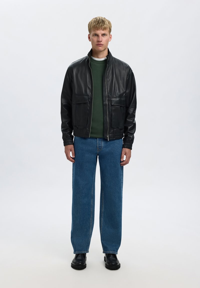 Lyshåret mand iført en sort læderjakke, grøn sweater, blå jeans og sorte sko står mod en ensfarvet lys baggrund.