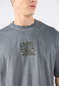 Graues Baumwoll-T-Shirt mit Rundhalsausschnitt, featuring besticktem Text "OUTER MIND VISIONS" in Dunkelgrün. Weiche Textur, kurze Ärmel.