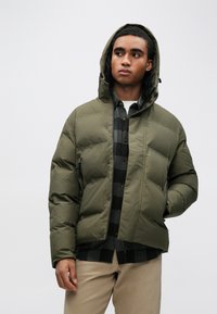 Olíva zöld kapucnis tollkabát cipzáros záródással, texturált anyaggal és feltűnő varrással; fekete és szürke kockás ing fölött rétegezve.