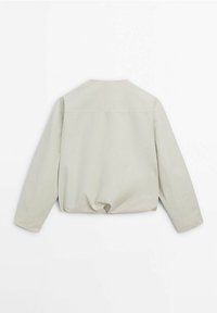 Beige langærmet jakke med lige snit, minimalistisk design og let samling i kanten, vist fra ryggen på en hvid baggrund.