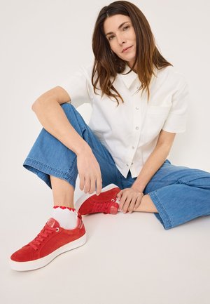Baskets en suède rouge avec semelles blanches, présentant un design à lacets et des éléments décoratifs. Portées avec des chaussettes blanches ornées de cœurs rouges.