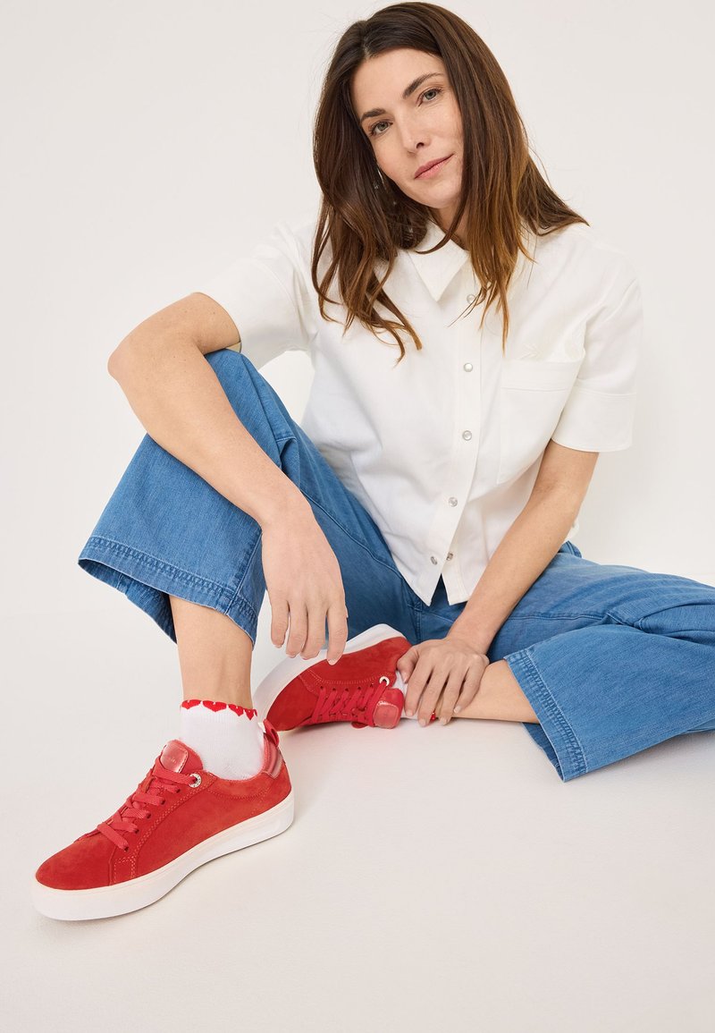 Sneakers rosse in suede con suole bianche, caratterizzati da un design con lacci e accessori decorativi. Indossati con calzini bianchi ornati da cuori rossi.