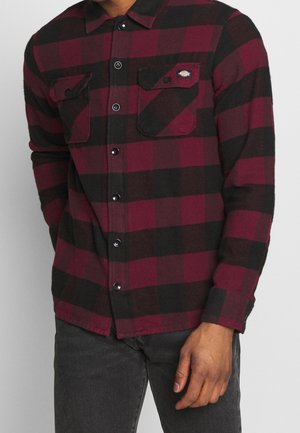 Homme portant une chemise en flanelle à carreaux rouges et noirs boutonnée avec deux poches poitrine et un jean gris foncé, main détendue sur le côté.