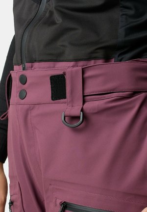 Close-up af taljeområdet, der viser mauve bukser med bæltestropper, sort D-ring, trykknapper, velcro-lap og lynlåslomme, båret med sort top.