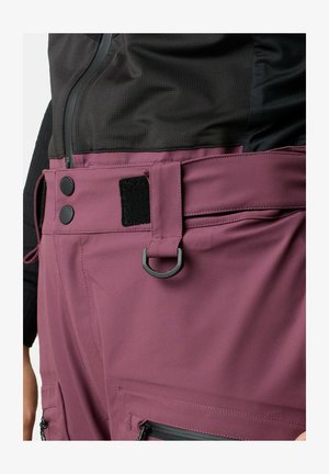 Close-up af taljeområdet, der viser mauve bukser med bæltestropper, sort D-ring, trykknapper, velcro-lap og lynlåslomme, båret med sort top.