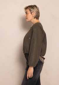 Cropped olive green zippered jacket in a textured fabric, featuring puffed sleeves and a simple pocket detail, paired with dark jeans.