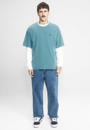 Mężczyzna z krótkimi ciemnymi włosami i wąsem, ubrany w turkusowy, oversize'owy t-shirt, białe długie rękawy, luźne niebieskie dżinsy i czarno-białe trampki.