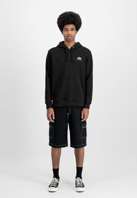 Felpa nera con cappuccio e tasca frontale, abbinata a pantaloni cargo neri al ginocchio. Presenta cuciture bianche e scarpe sportive casual.